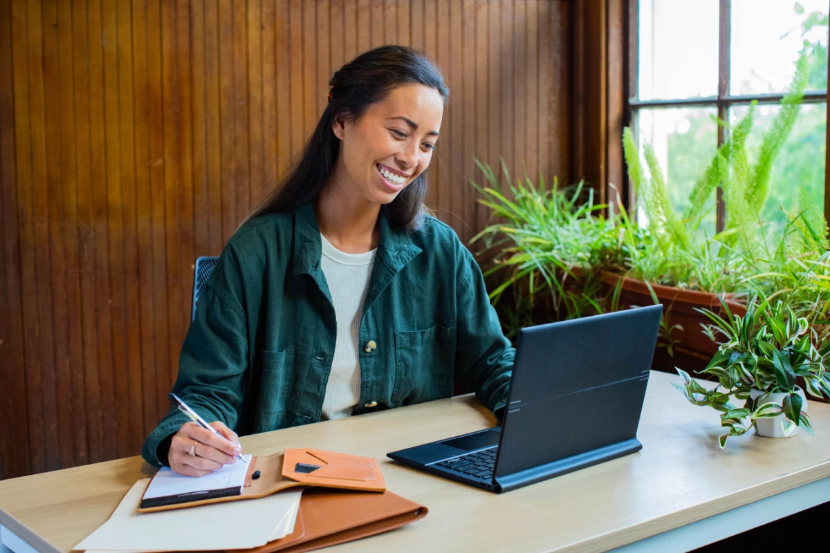 Hybrid work. Woman sitting at casual desk at home with an HP Elite Folio which is a commercial device. Taking notes and smiling. Windows 11 Pro.