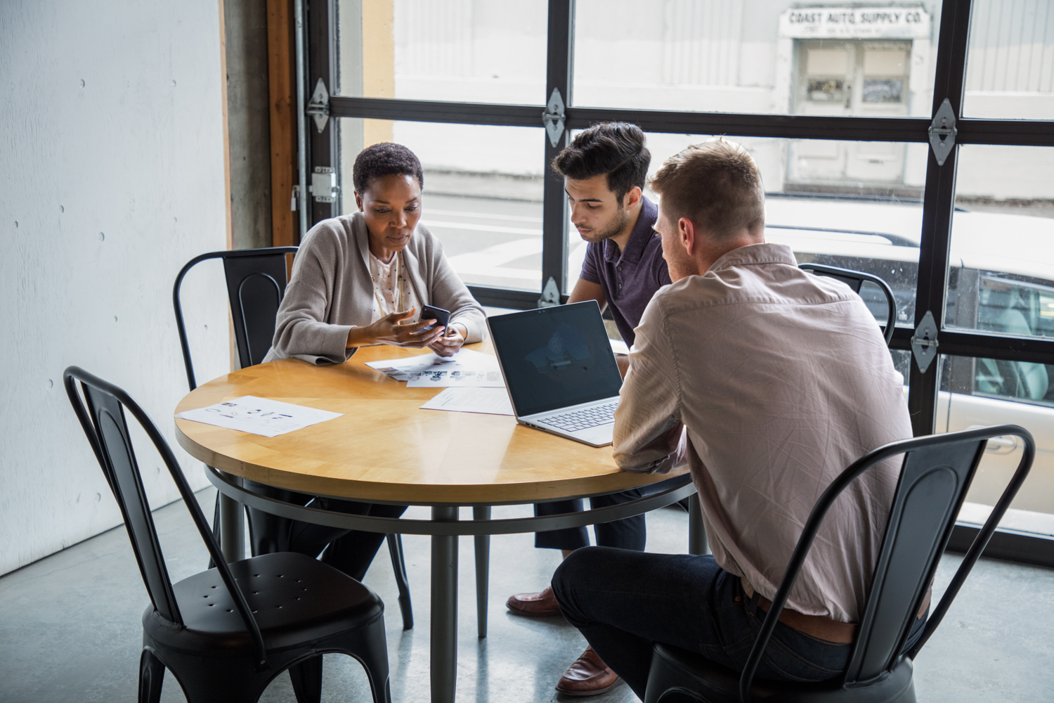 One female and two male workers sitting at round table in casual office setting or caf&eacute;/coffeehouse. The woman is holding a phone to show something to one of the men. The other man is using a Surface Laptop.