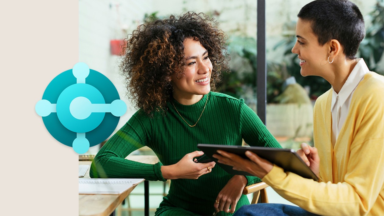 A woman in a green shirt talking to a woman in a yellow shirt who's holding a tablet. The Dynamics 365 Business Central icon is on the left side of the image.