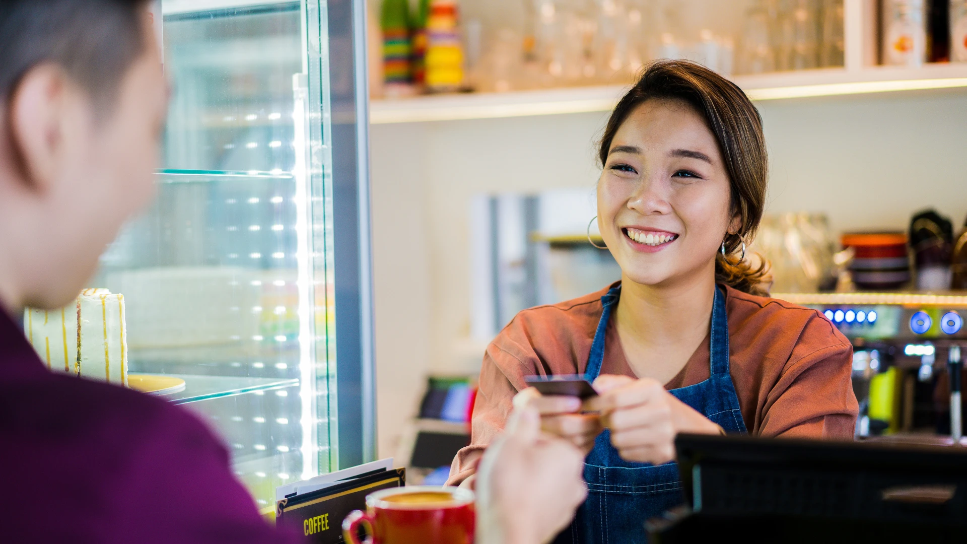 Person at a caf&eacute; counter holding a payment card during a customer transaction, with a coffee machine and display case visible in the background
