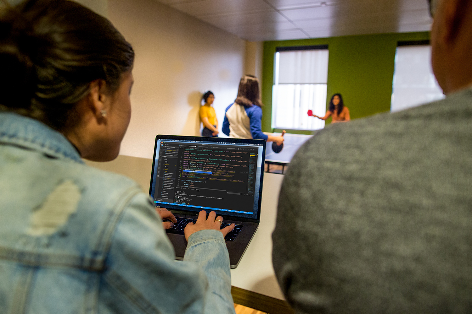 A college student works on a laptop, while fellow students play table tennis in the background.