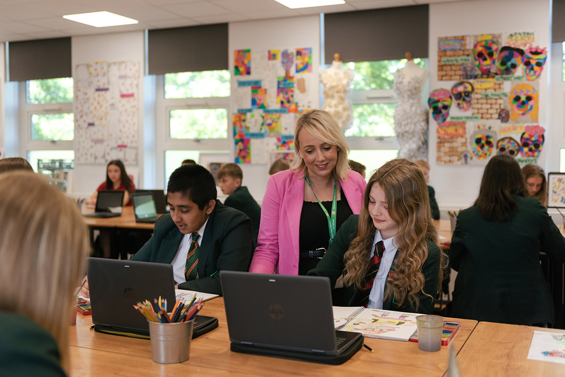 A high school teacher looks over the shoulder of a student working on a laptop.