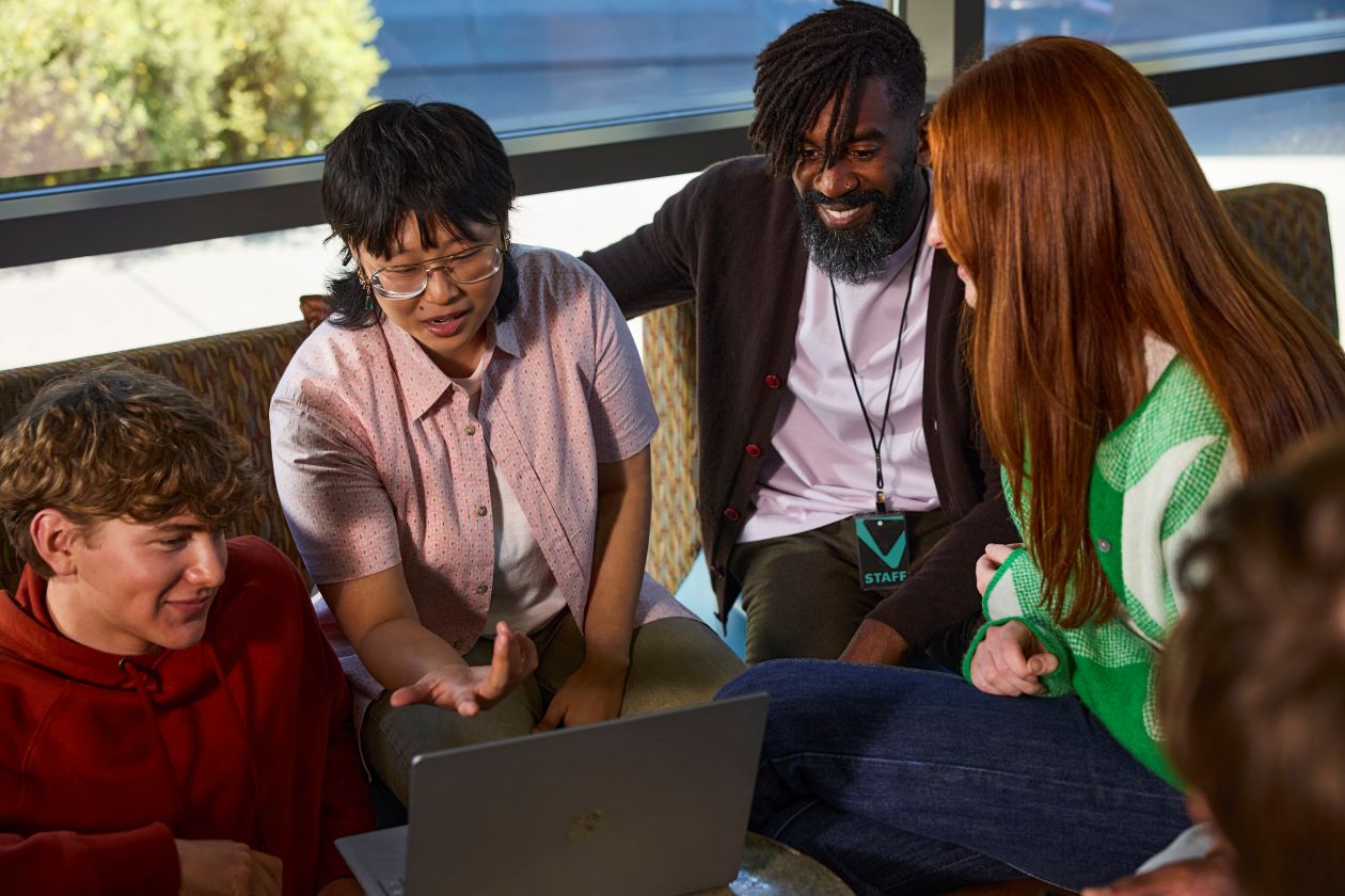 An instructor and a group of students collaborate on a laptop in a common area at a college.