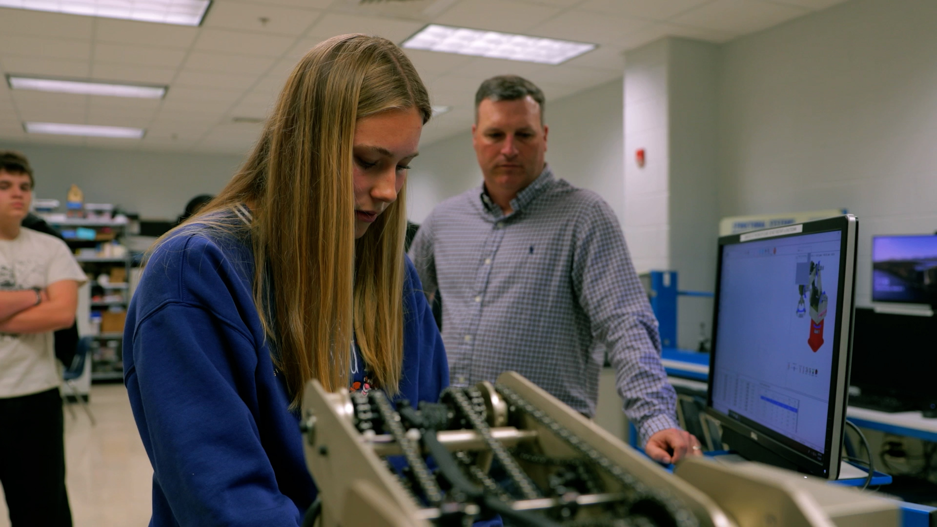 A technical classroom scene showing a student working on a robotic component from plans on a screen. A teacher and additional students look on from behind her