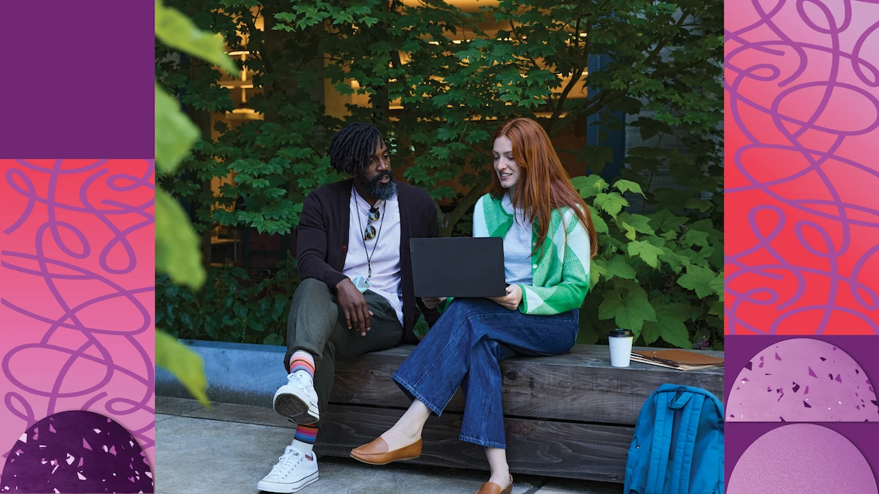 Two people sit together on a bench surrounded by trees and look at the screen of a laptop