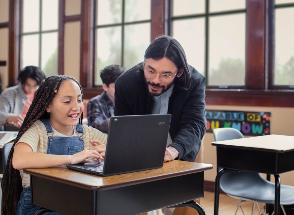 An educator in a classroom helps a student with schoolwork on her laptop.