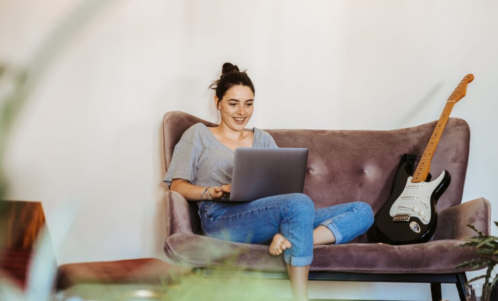 A person sitting on a sofa using a laptop. There’s a guitar nearby.