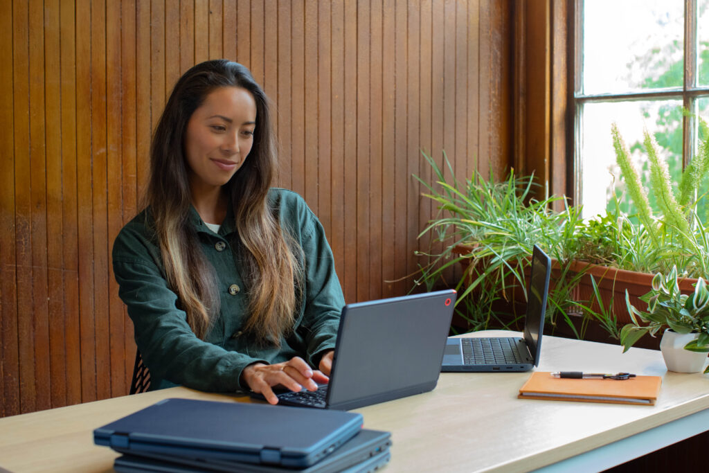 A person sitting at a table working on a laptop, with another laptop and tablets on the table. 