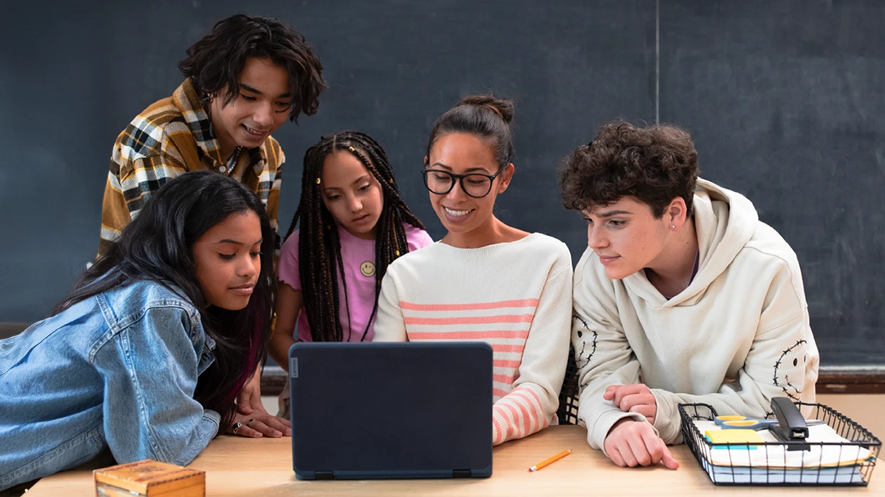 An educator and four students looking at a laptop at the educator&rsquo;s desk in a school classroom