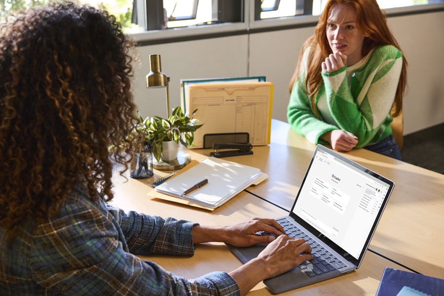 An educator sits across from a student at a desk in a school office and uses Copilot Chat on a laptop.