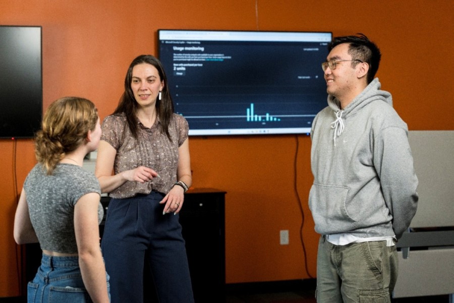 An IT professional and two students stand in front of a screen showing a cybersecurity dashboard in a university office.