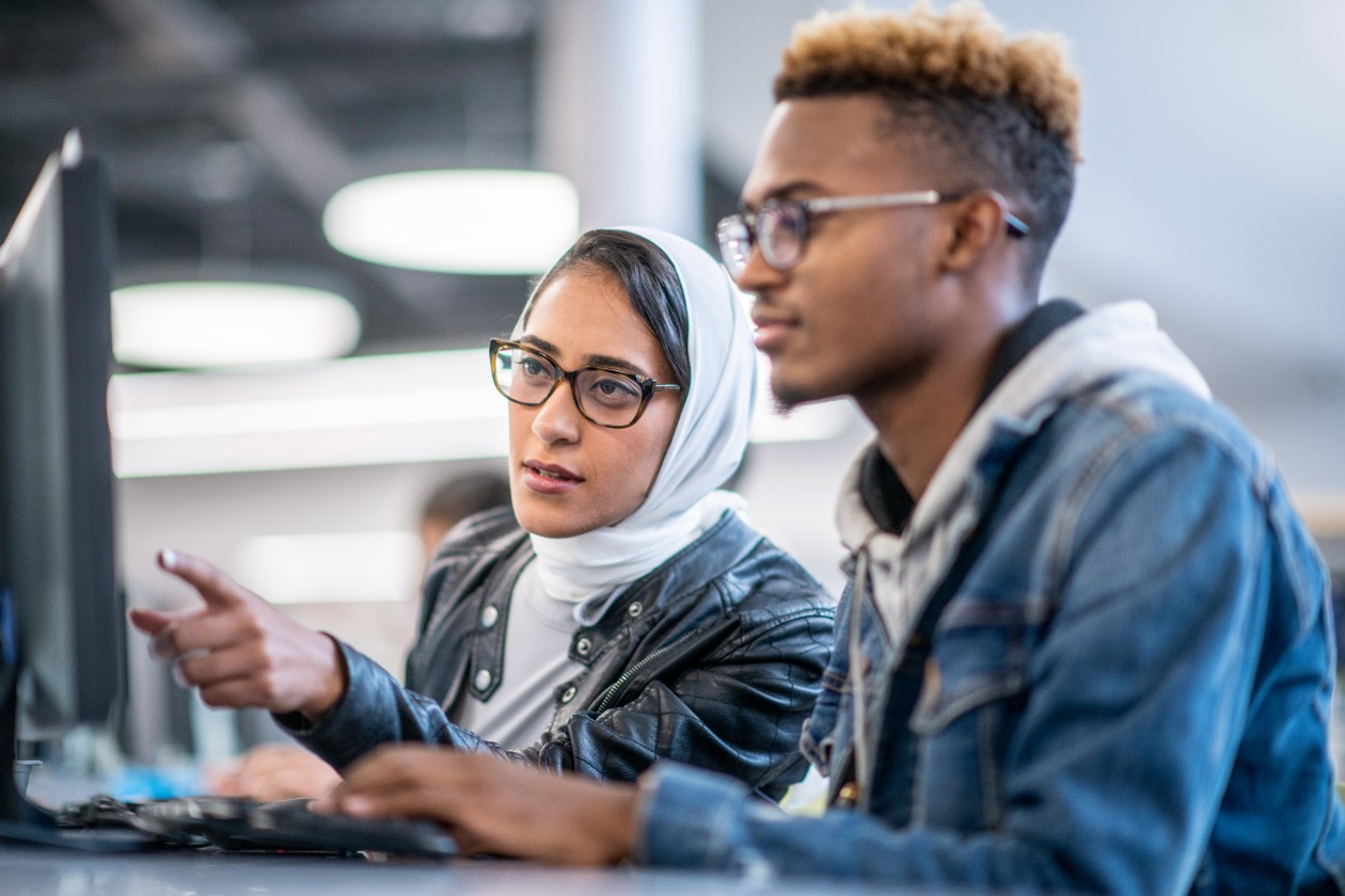Two higher education students sit at a desk and work on a computer.