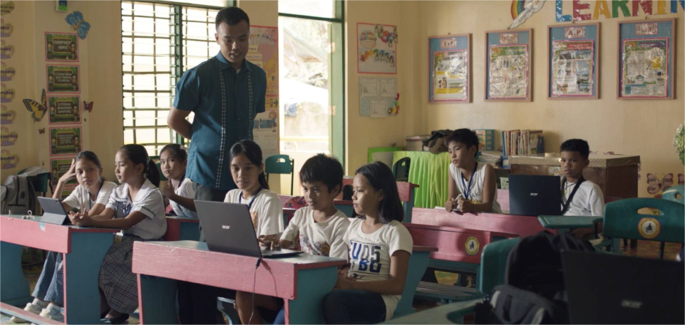 Teacher walking between two rows of desks as students work on laptops in pairs or groups of three.