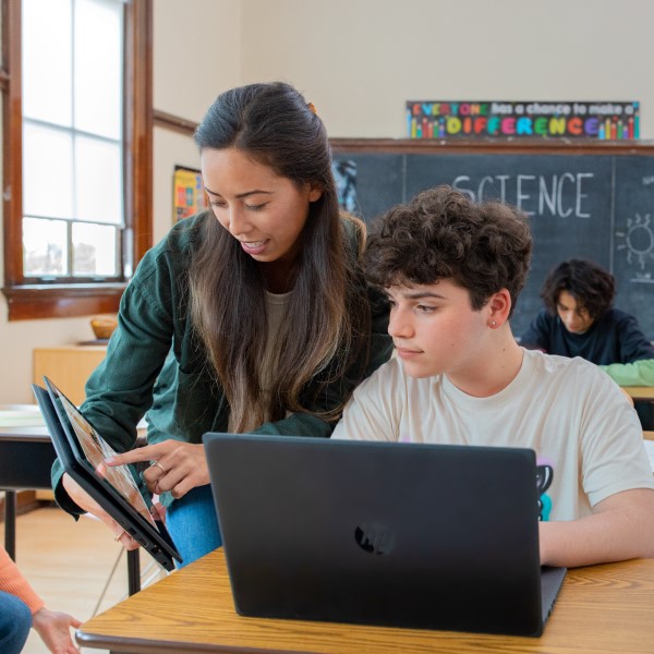 Decorative. A teacher engages with a student at his desk. They're both using laptops.
