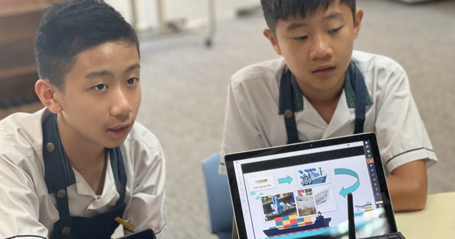 Two students sit at a table with a computer facing away from them. The computer screen shows a chart with a ship.