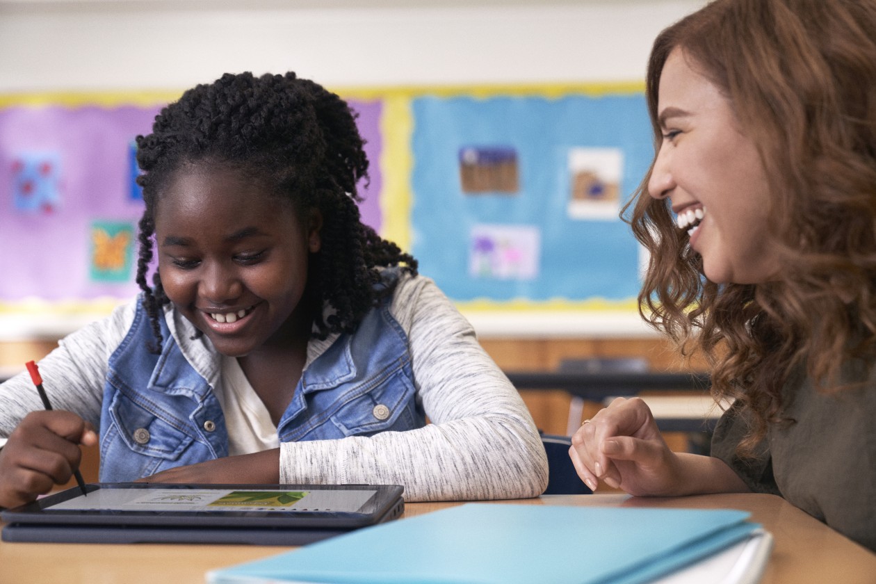 A student uses a digital pen to write on a laptop while an educator smiles encouragingly.