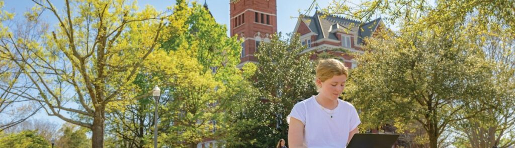 A student sits and uses a laptop outside on a college campus.