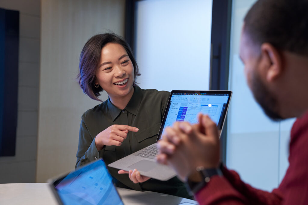 Two colleagues collaborate at a meeting table, with one smiling and pointing to data on a Microsoft Surface laptop screen while the other listens.