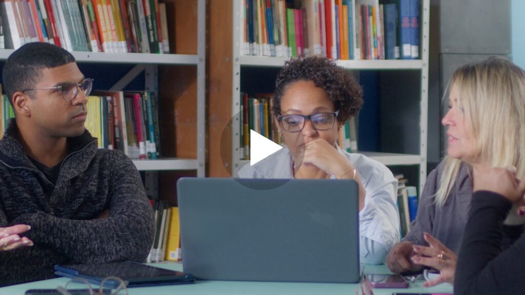 Three people sitting at a table in a library looking at a computer.