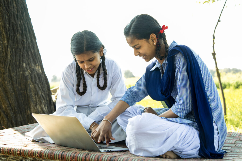 Two young female students in school uniforms sit together outdoors on a woven mat beneath a tree, smiling as they work on a laptop with a green field visible in the background.