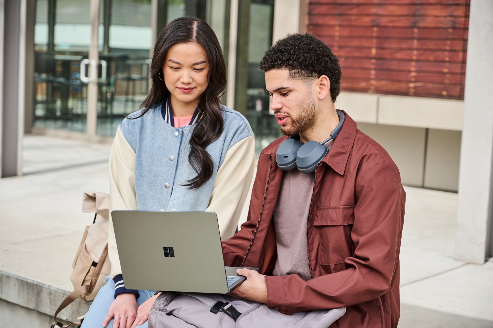 Photograph of a male and female sitting outside of a brick building. The male is holding a Surface Laptop 13 Ocean device