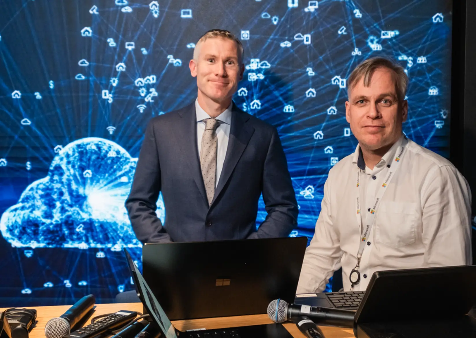 Conor Kelly standing at a desk wearing a blue suit, with Henning Kilset sitting at the desk wearing a long sleeve collared shirt.