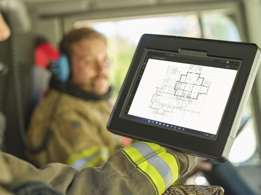 In the forefront, the arm of a firefighter sitting in the passenger seat of a firetruck holding a Surface tablet to look at blueprints of a building and a male firefighter in the background in the driver’s seat with a headset on.