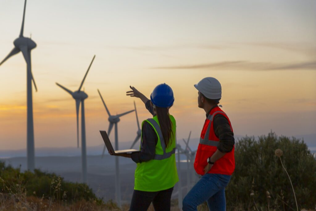 One woman, holding a laptop, and one man, working in a wind turbine at sunset.