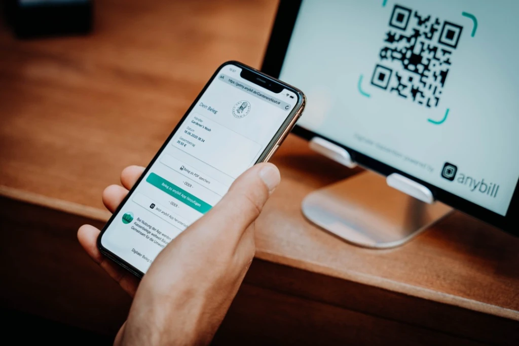 Auf dem Bildschirm eines Smartphones sieht man einen Online-Beleg. Im Hintergrund sieht man einen QR-Code, der zu dem Beleg führt, auf einem Tablet.