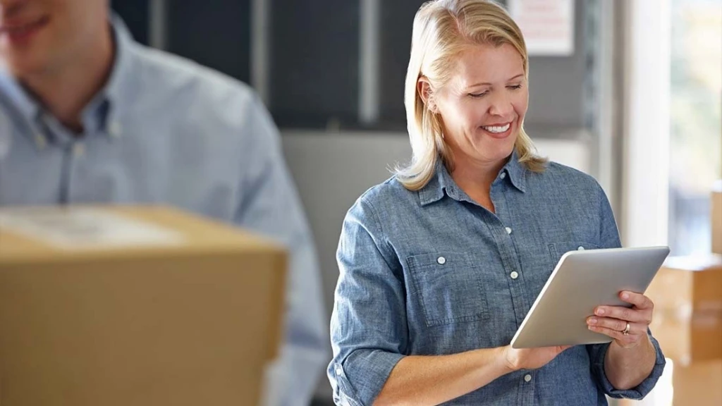 Eine Frau, umgeben von Paketen, blickt auf einen Tablet Computer.