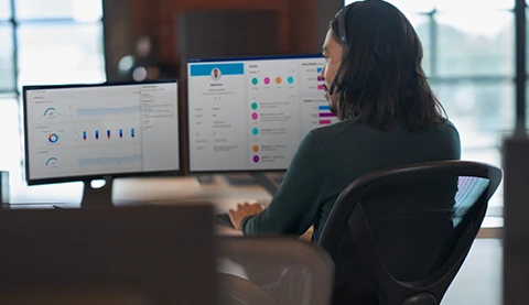 Frau sitzt an einem Schreibtisch und blickt auf zwei Bildschirme mit Diagrammen.