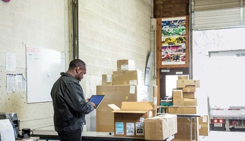 Ein Lagerarbeiter vor vielen Paketen, er hält ein mobiles Device in den Händen 