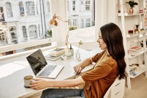 A woman is drinking coffee while using her Lenovo Yoga while sitting at her desk using Windows Virtual Desktop in her bright sun-filled apartment.
