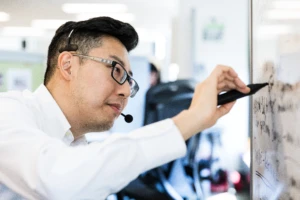 Male worker writing a cloud strategy with black pen on whiteboard in financial office. He is wearing a phone headset and glasses.