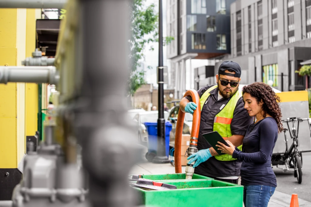 Two water frontline workers in a city. One is holding a hose, the second is holding a tablet that they are both looking at.