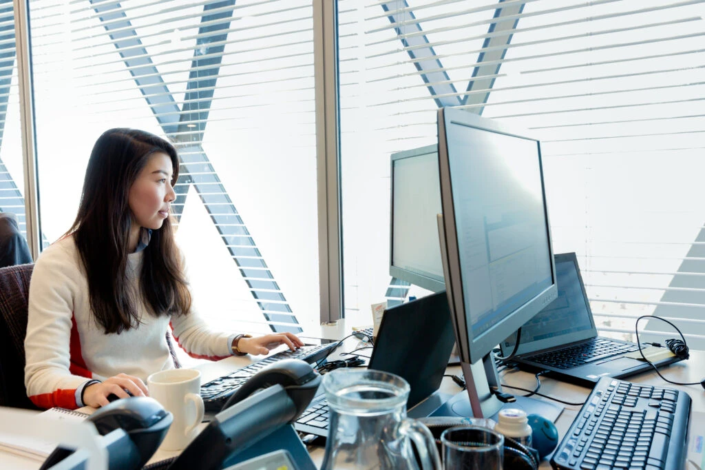 Female office worker at workstation near window, looking at desktop monitor (screen not shown). Open laptop also on desk (screen not shown).