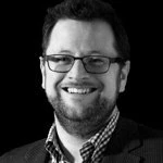 A man smiles at the camera. He has glasses, dark hair and stubble.