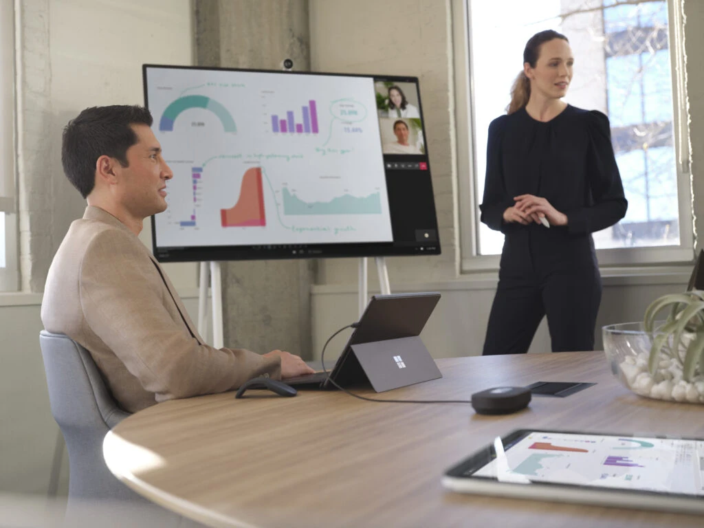 Two adults conducting a meeting in a conference room while collaborating over a Microsoft Teams video call presented on a Surface Hub 50&rdquo; device. Two Surface Pro 8 devices are also shown. One in laptop mode with Surface Pro Signature Keyboard and one in tablet mode with Microsoft PowerBI shown.