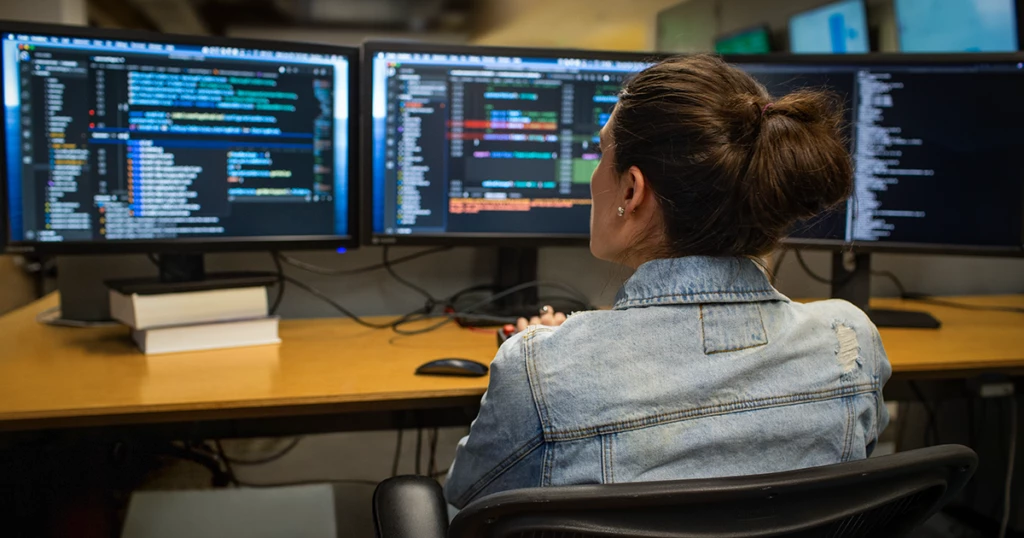 Real people, real offices. Developer coding at their workspace in an enterprise office, using Visual Studio on a multimonitor setup.