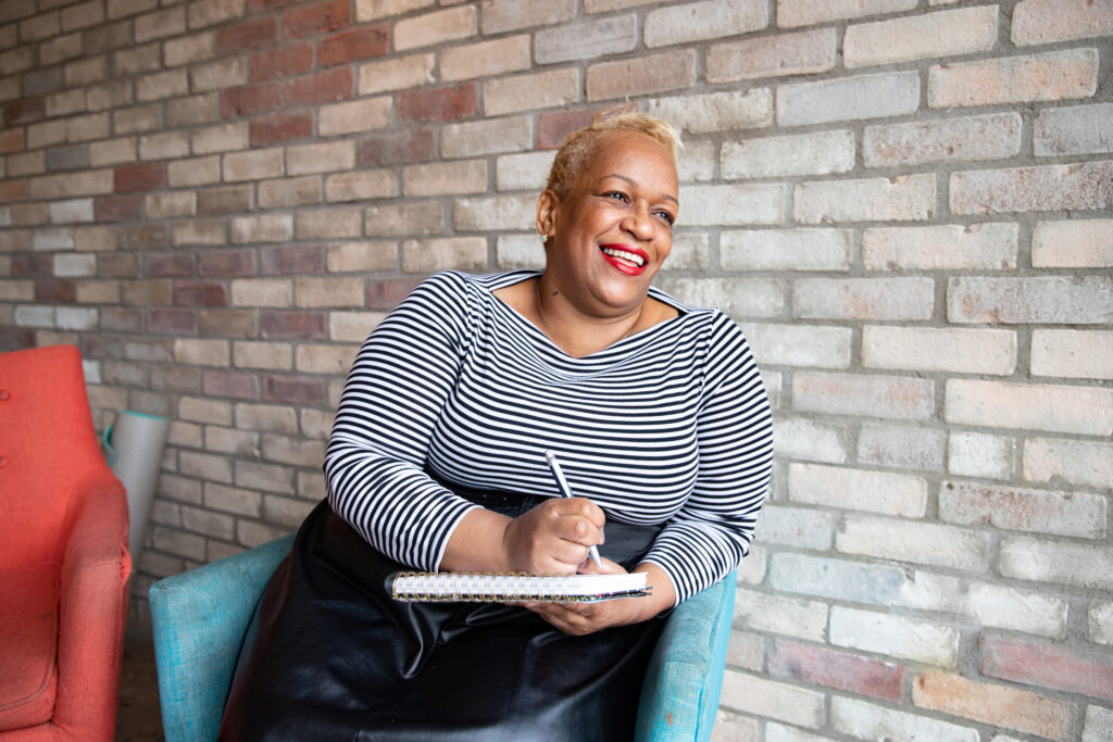 An mature black entrepreneur holding a notebook sits in an armchair against a brick wall in a cafe.