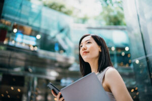 Successful and modern young Asian businesswoman carrying smartphone and laptop, commuting to work in central business district against contemporary corporate buildings in the city. Female leadership. Business on the go