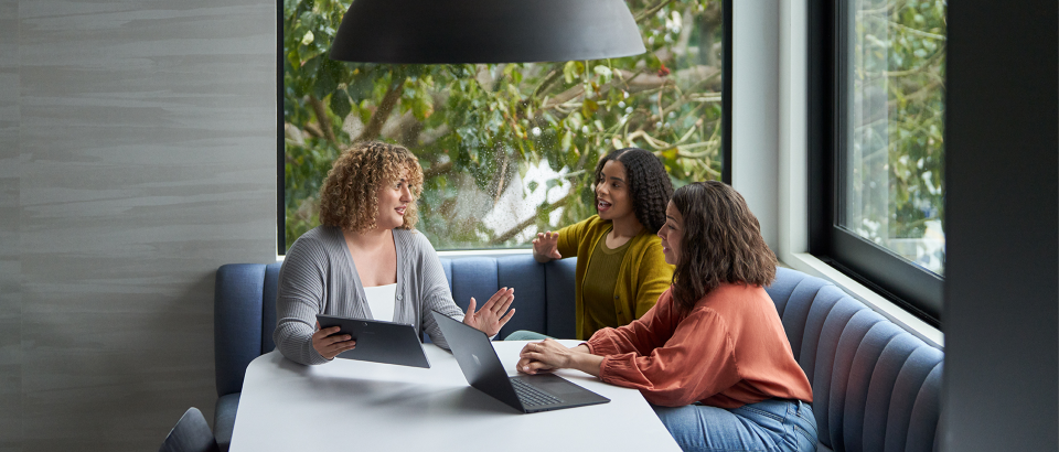 Three people dressed in casual sweaters engaged in discussion with 2 laptops open on a table in front of them.