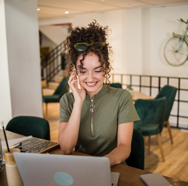 A woman talks on the phone while working on a laptop in a cafe.
