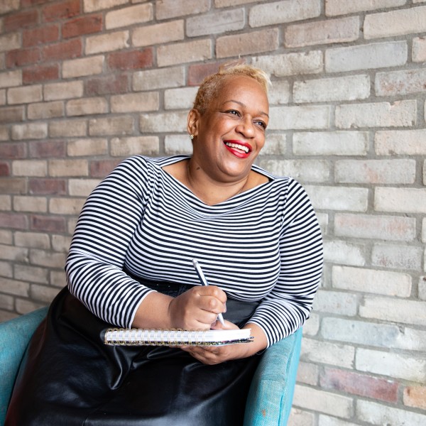 A woman sitting in a chair, smiling, holding a notebook and pen.