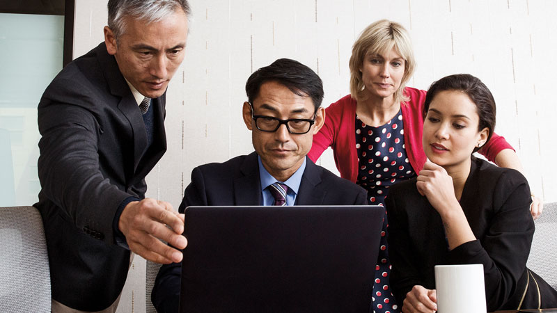 Four people sitting and standing at a desk analyzing a laptop computer