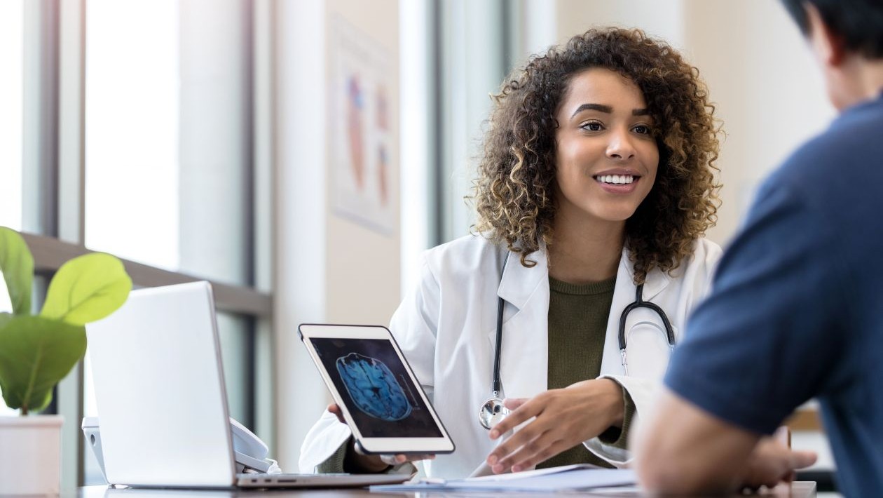 A woman in a white coat holding a tablet talking to a man