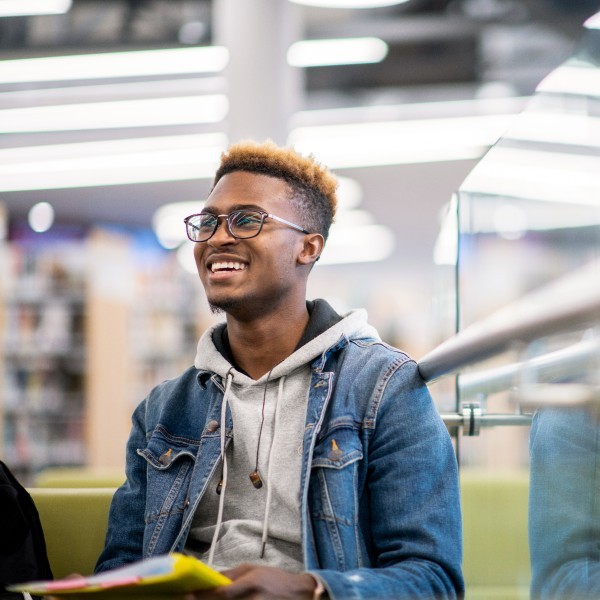A smiling male university student wearing a hoodie and jean jacket sits in a brightly lit library.