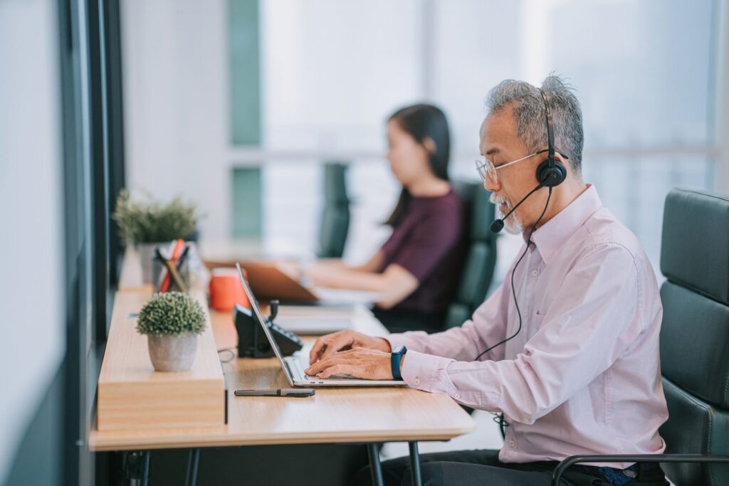 Person wearing a headset uses a laptop at a desk in an office workspace while another person works at a nearby desk in the background.