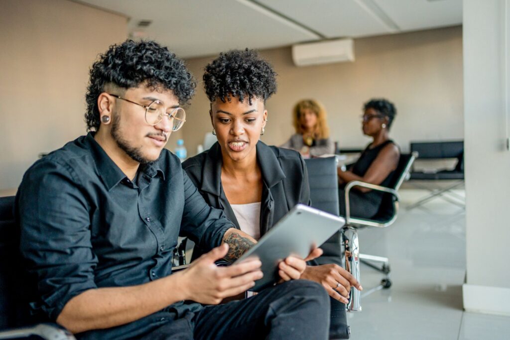 Two people review information on a tablet while seated in a meeting room, with additional attendees in the background.