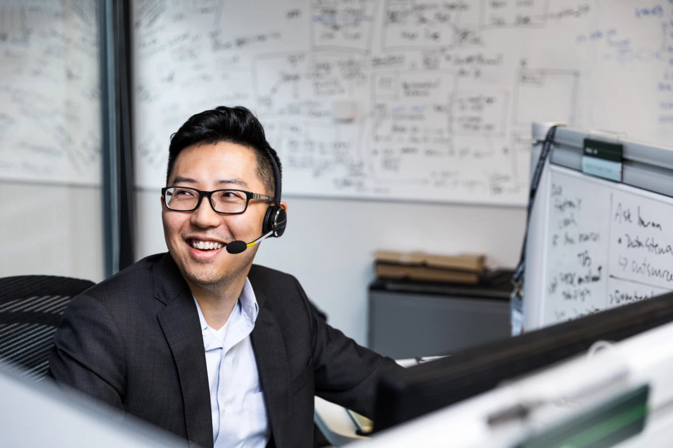 Male worker sitting at cubicle desk in financial office, wearing glasses and phone headset. He is smiling and looking up to his right. A large whiteboard covered in black written text is on wall behind him. A smaller whiteboard also partially visible on his desk‘s cubicle wall.